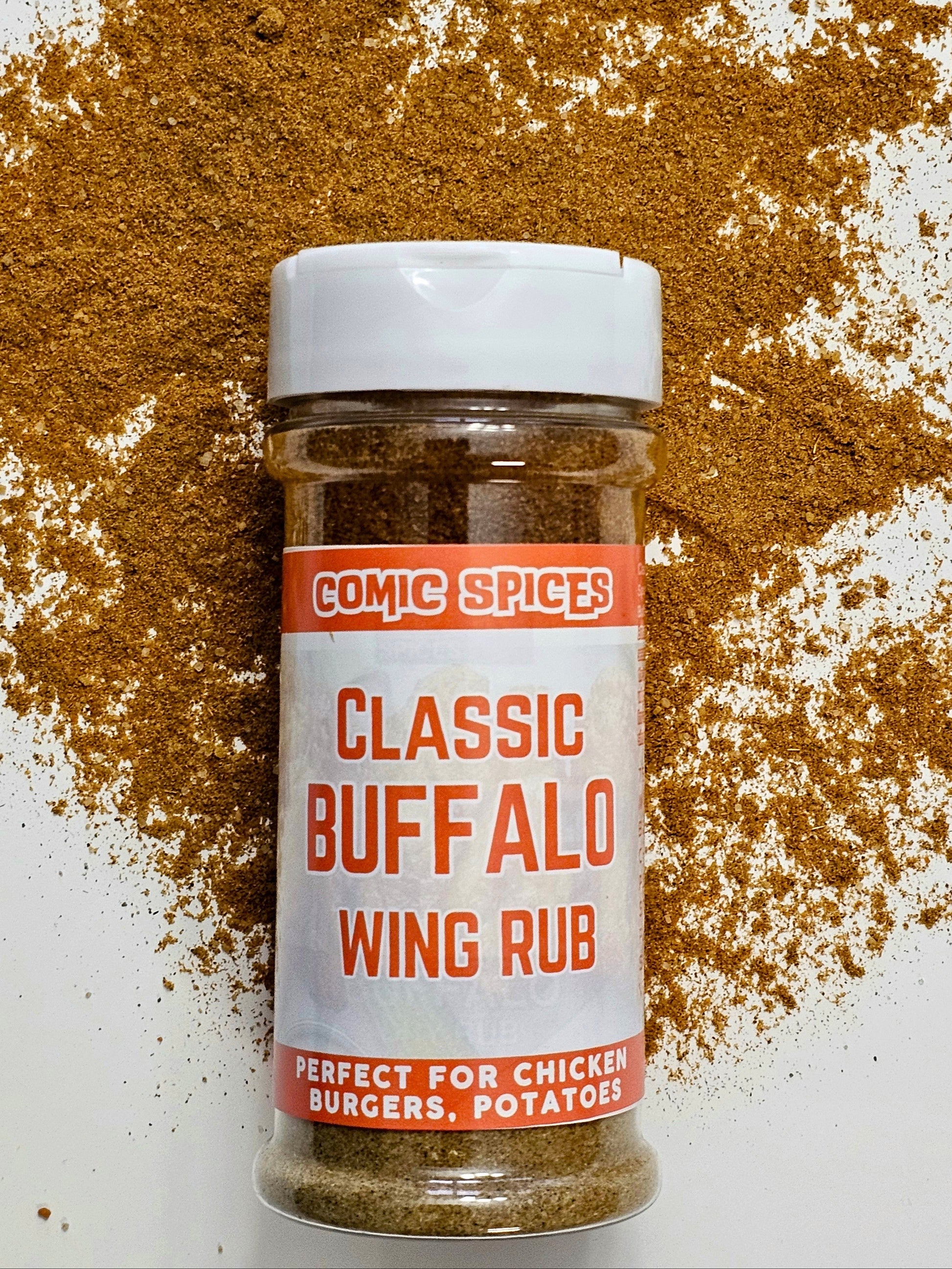 spice jar labeled Comic Spices Classic Buffalo Wing Rub lies on its side, surrounded by a generous amount of the reddish-brown spice blend scattered on a white surface. The label highlights that the rub is perfect for chicken, burgers, and potatoes. The jar has a white flip-top lid